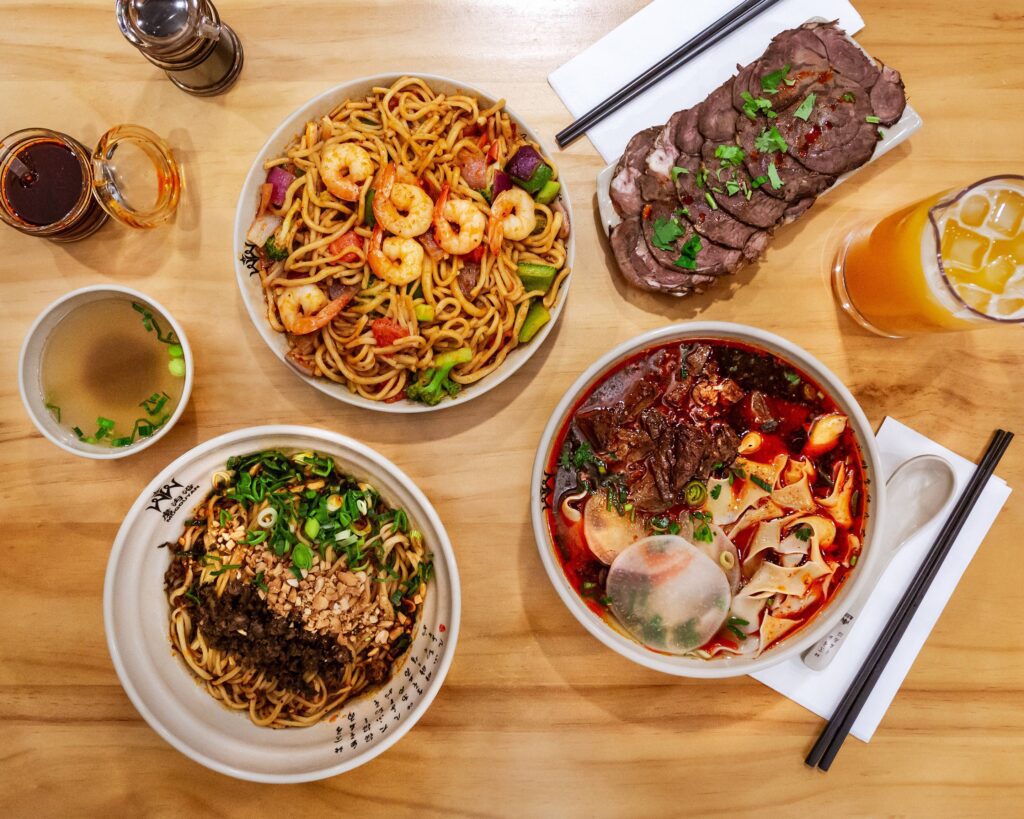 overhead shot of several hand pulled noodle dishes, steak and a drink