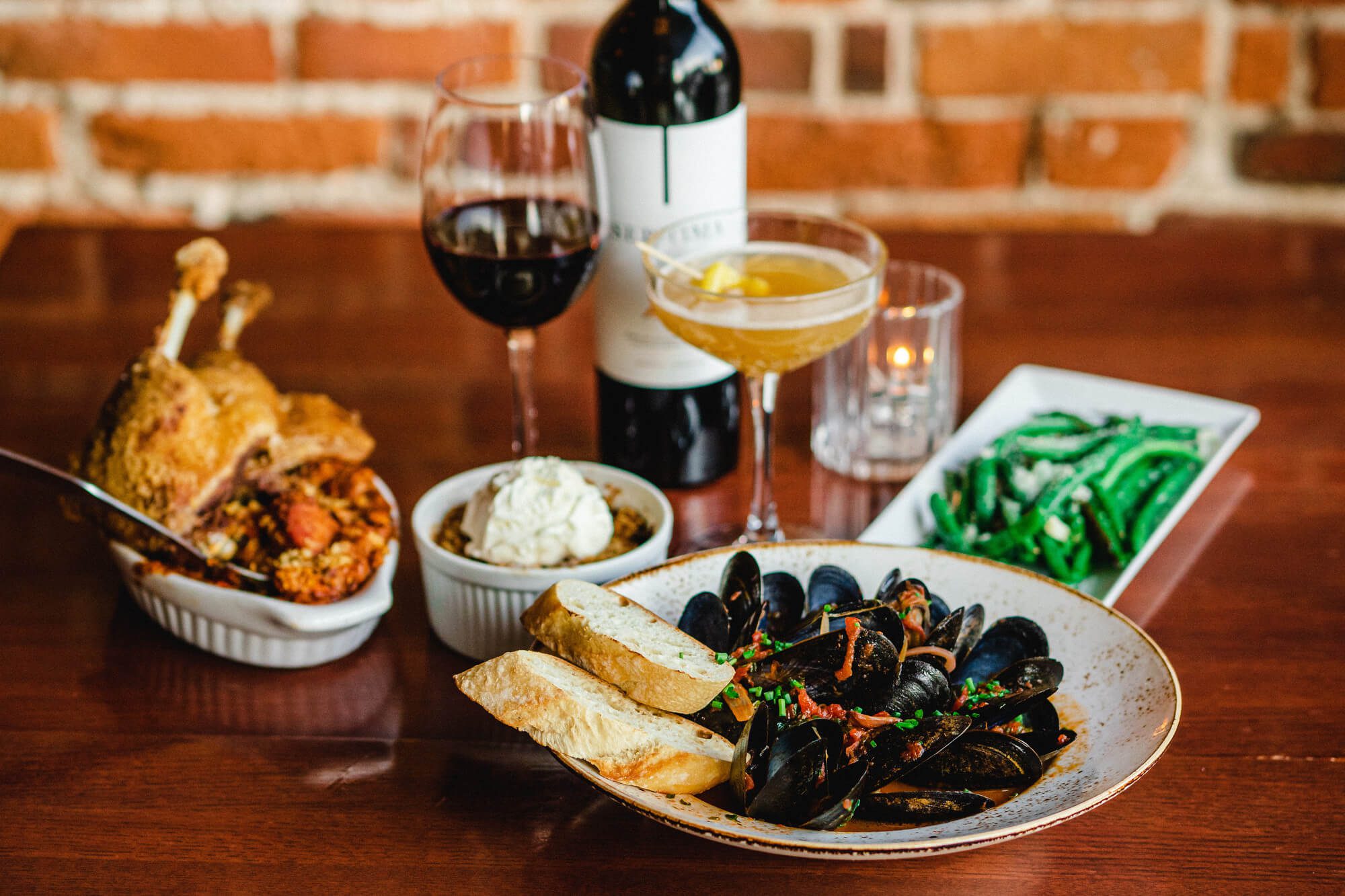 bottle of red wine, glsas of red wine, yellow cocktail, tealight candle, green beans, mussels and baguette and chicken dish on a table against an exposed brick wall at Black Dog Tavern