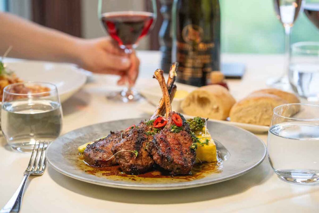 Shot of lamb rack at Peninsula Estate Winery with a hand holding on to the stem of a glass of red wine int he background 