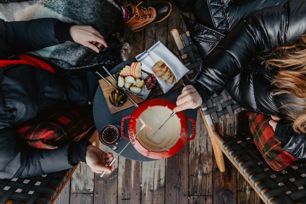 overhead shot of a fondue spread in a big red fondue pot with two hands reaching for various items