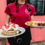 girl with braids in red shirt holding a plate of fluffy pancakes topped with fruit and whipped cream in one hand and homefries and sausage in the other