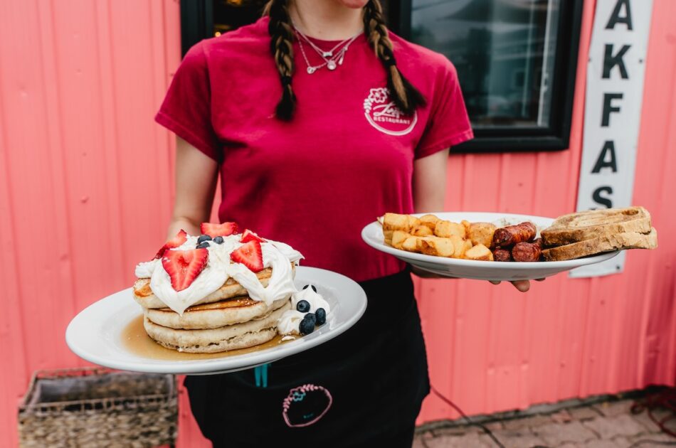 girl with braids in red shirt holding a plate of fluffy pancakes topped with fruit and whipped cream in one hand and homefries and sausage in the other