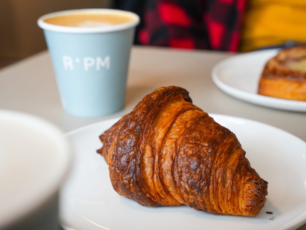 close up of a croissant and an RPM Bakehouse cappucino in a blue come behind that