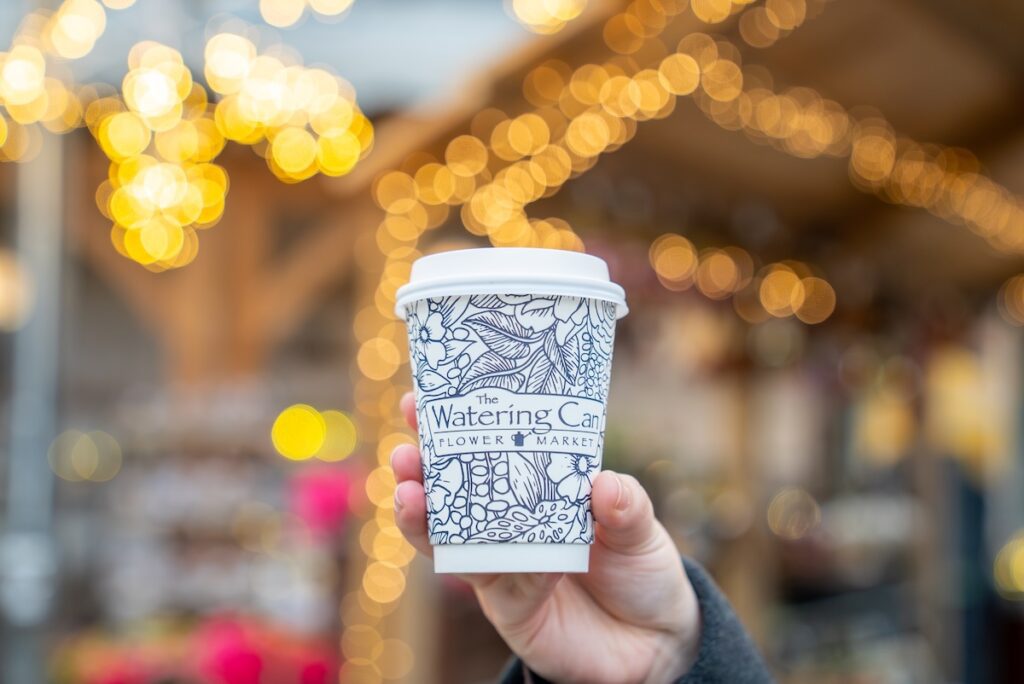 hand holding Watering Can Flower Market coffee cup with twinkle lights blurred in the background