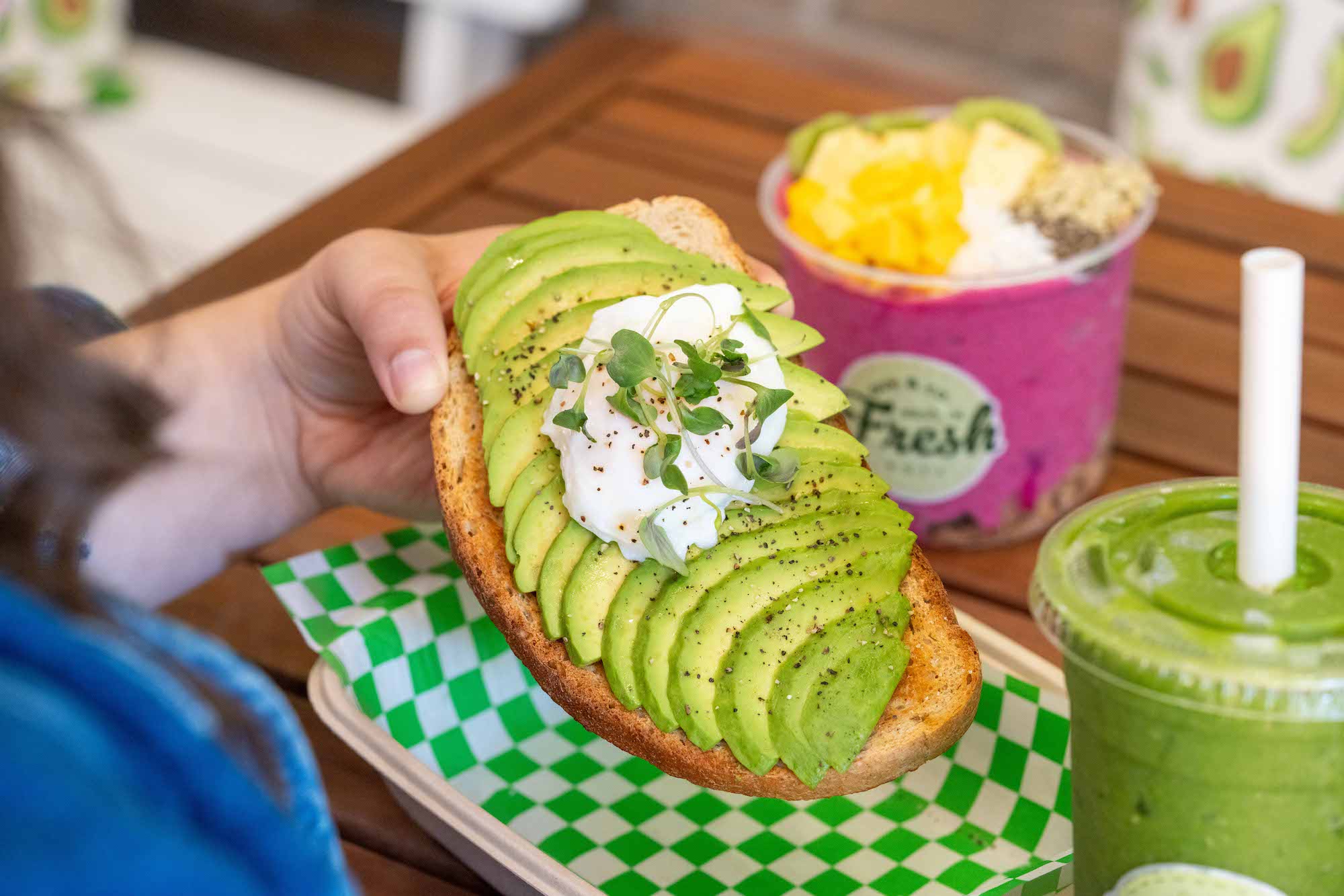 close up shot of avocado toast with a poached egg and micro green on top. In the background is a brightly-coloured pink fruit parfait and a bright green smoothie.