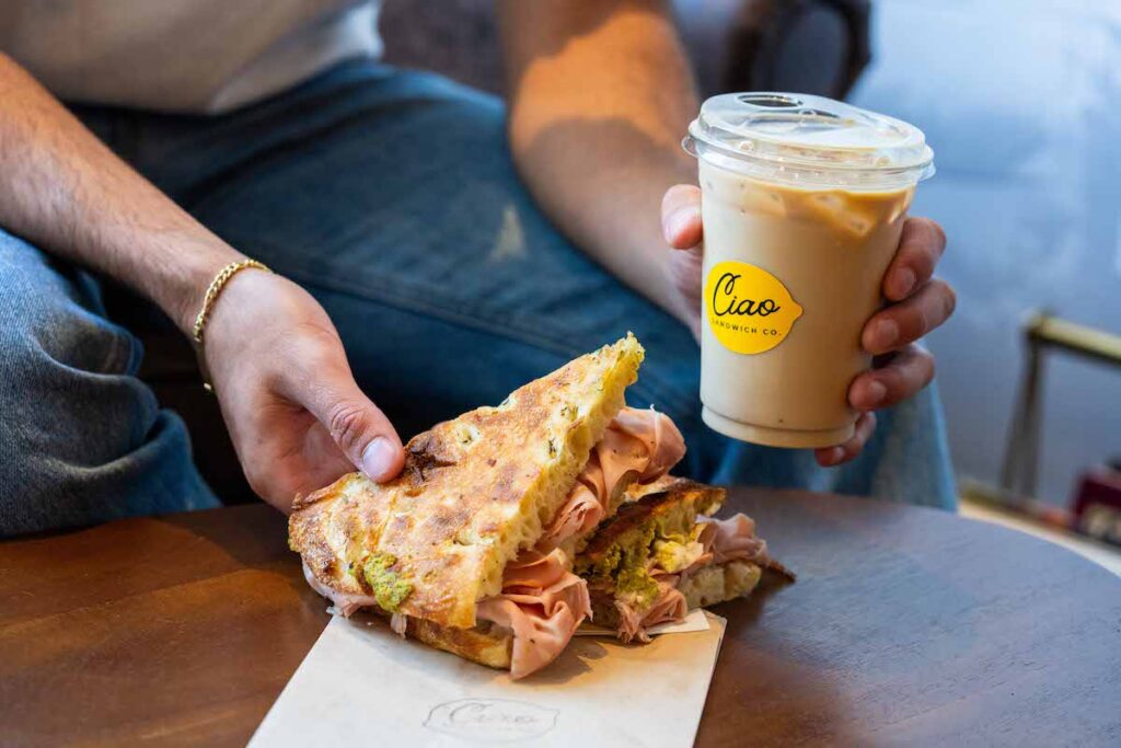 hands holding a large mortadella sandwich on foccacia and an iced coffee