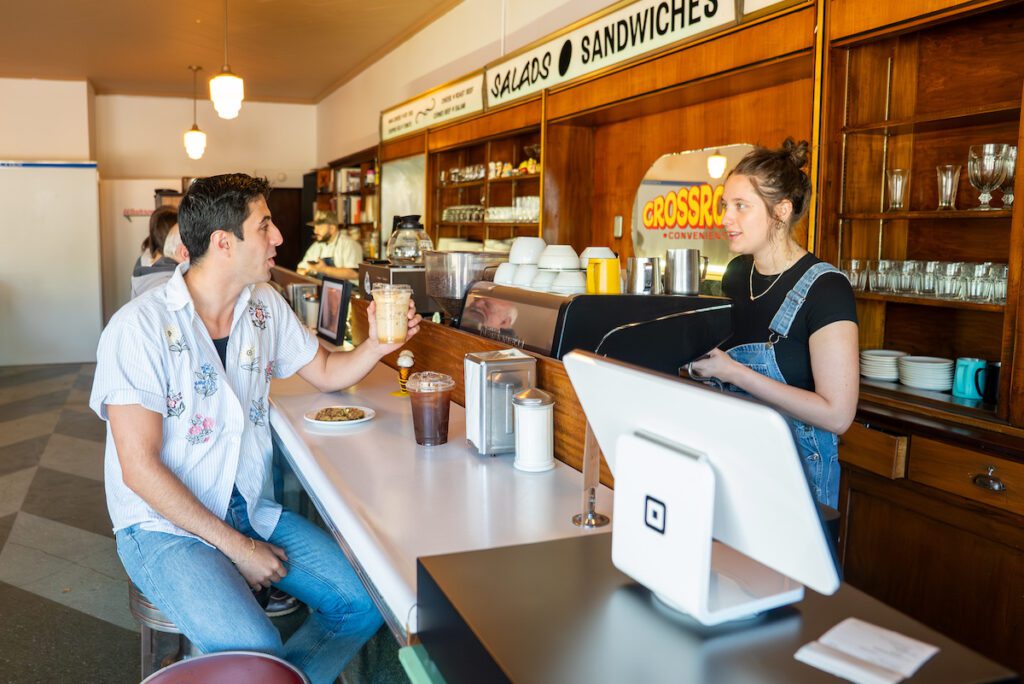 Zain with a cookie and an iced latte at the counter talking to person working at Crossroads Convenience