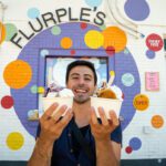 Zain holding up two bowls of ice cream at Flurple's in front of colourful mural