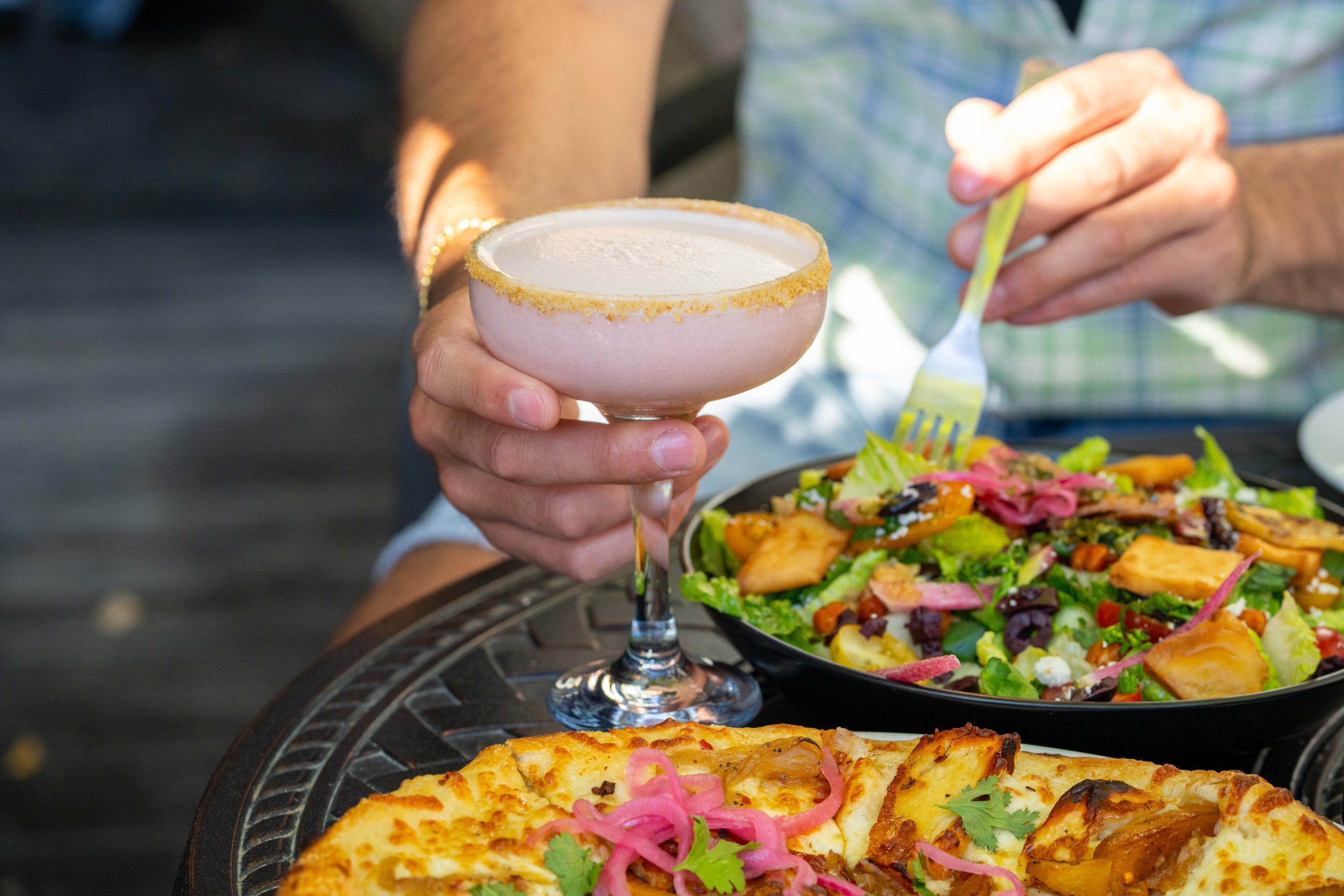 close up of a hand holding a purple cocktail over a brightly coloured salad and wood-fired pizza