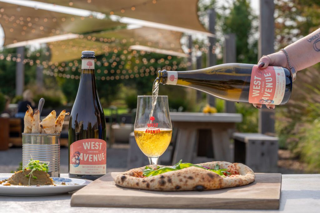 cider being poured into a West Avenue glass behidn wood fired pizza
