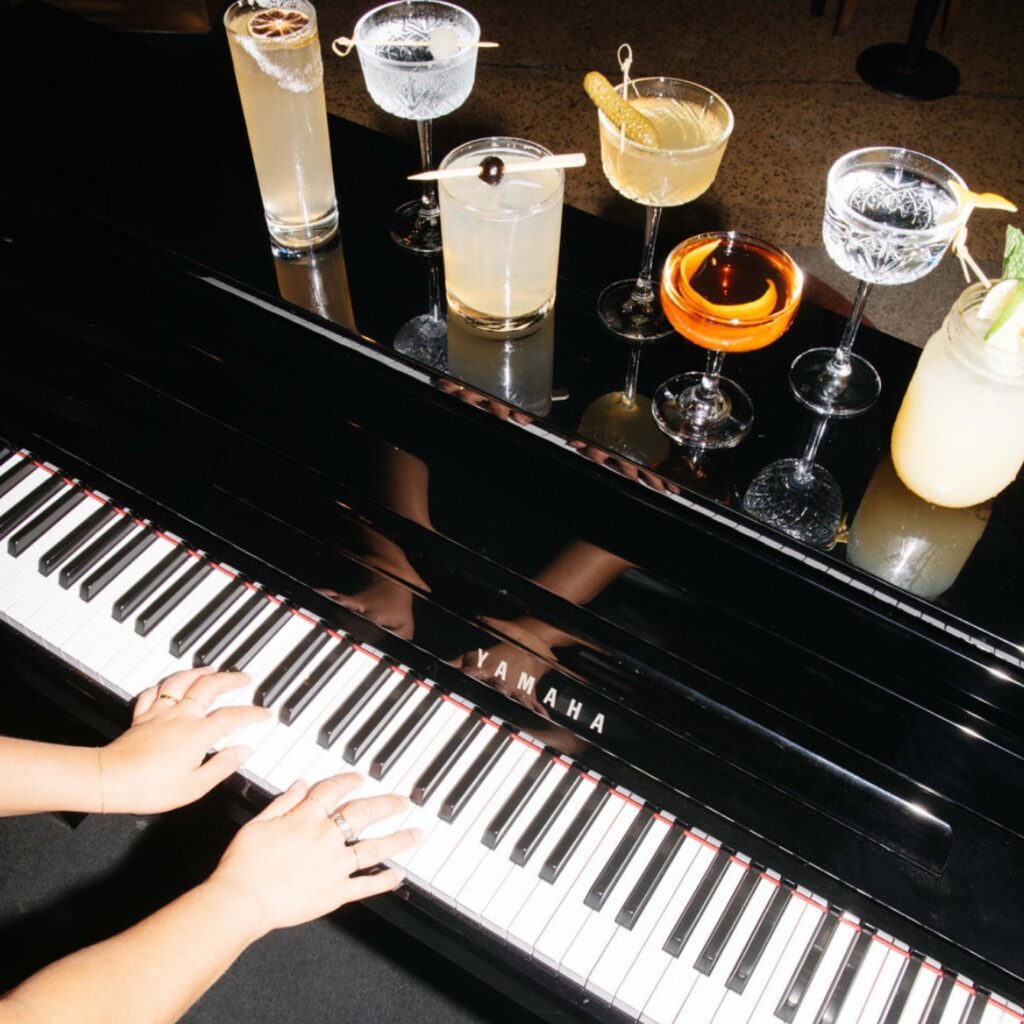 An overhead shot of two hands playing a piano with seven cocktails on it