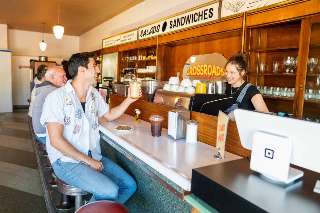 Zain with a cookie and an iced latte at the counter talking to person working at Crossroads Convenience