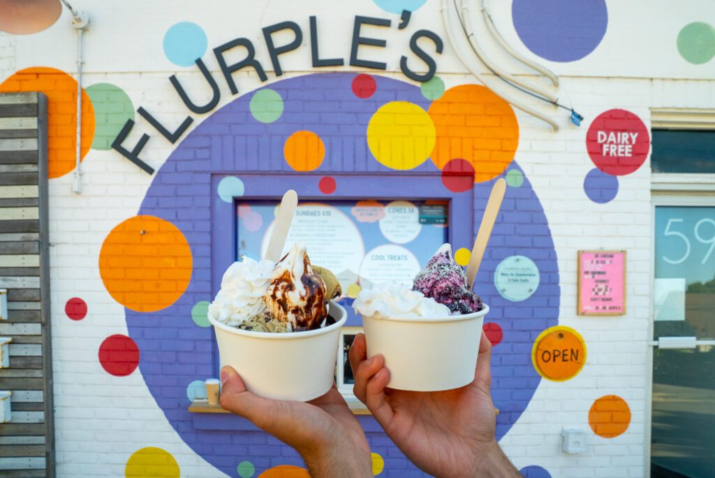 Two hands holding cups of vegan soft serve topped with whipped cream and sauce in front of the colourful Flurples dairy-free ice cream shop in Sudbury.