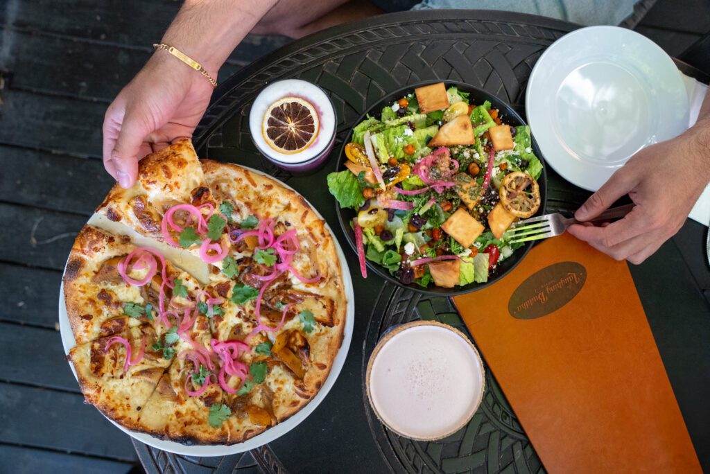 Hand holding a slice of pizza topped with pickled onions and cilantro next to a colourful salad and cocktails at The Laughing Buddha in Sudbury.