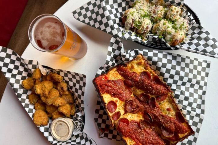 table shot showing a glass of beer, fried coins of pickles, round croquettes topped with melted white cheese, and pizza topped with red pepperoni