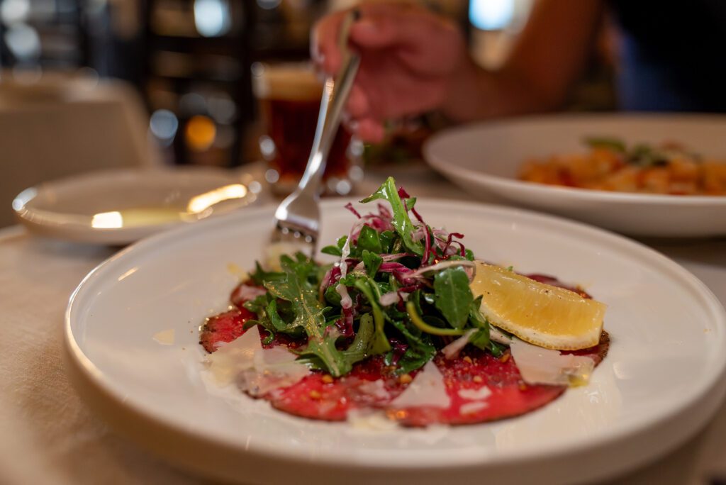 Thinly sliced beef carpaccio topped with arugula, shaved cheese and a drizzle of olive oil on a white plate.