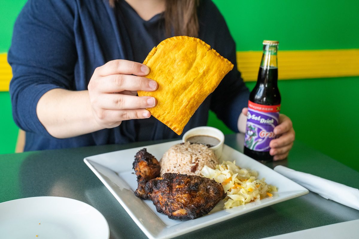 close up of hands holding a Jamaican cola and pattie in front of a plate of jerked chicken, jollof rice and more