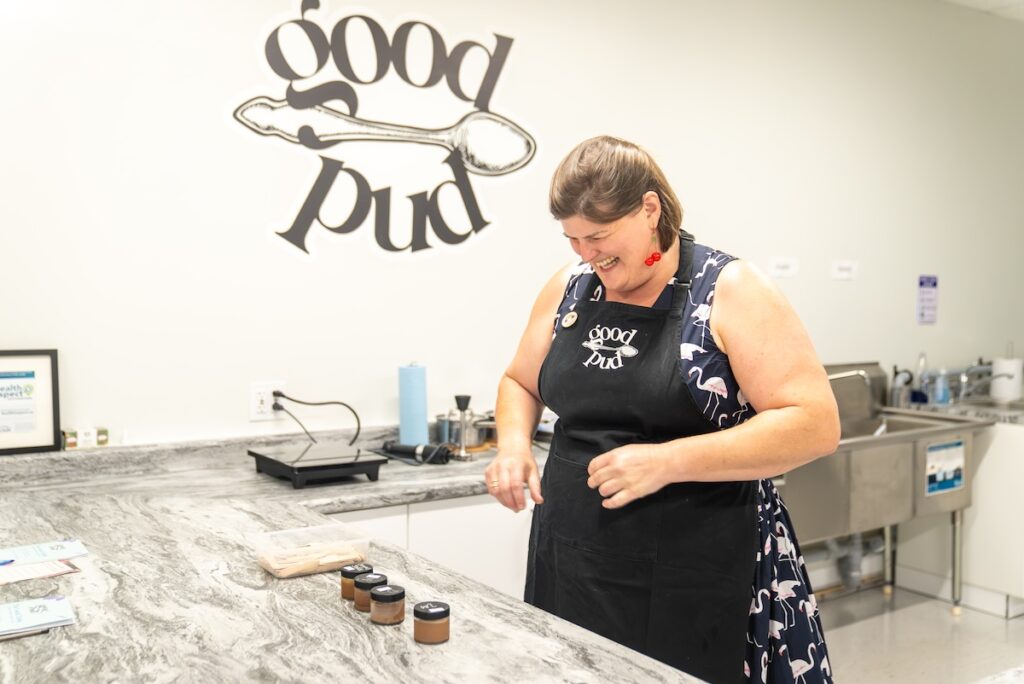 Woman smiling and laughing in front of pudding line up wearing a GoodPud apron